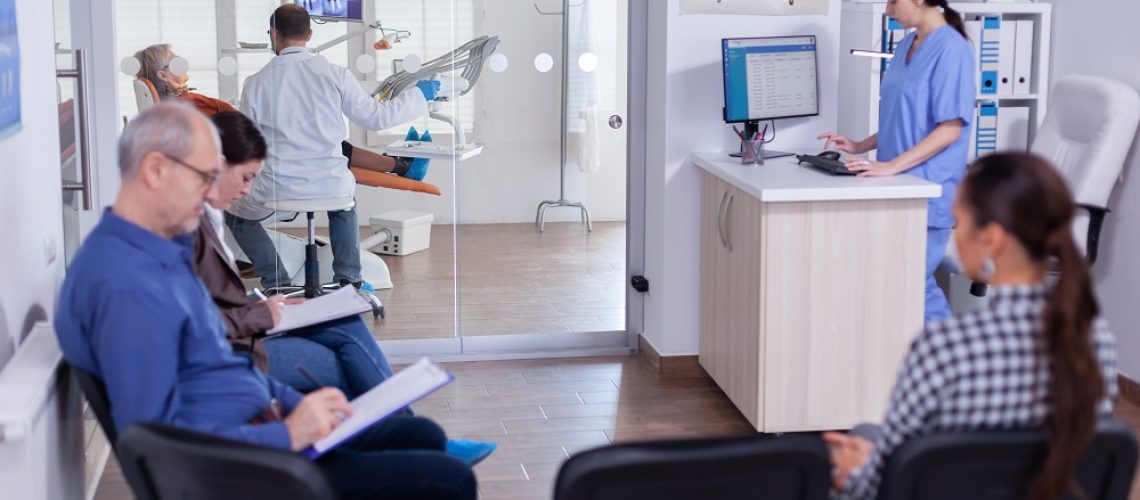 Crowded stomatology waiting area with people filling form for dental consultation. Stomatoloy specialist denstiry treating senior woman cavity. Receptionist working on computer.